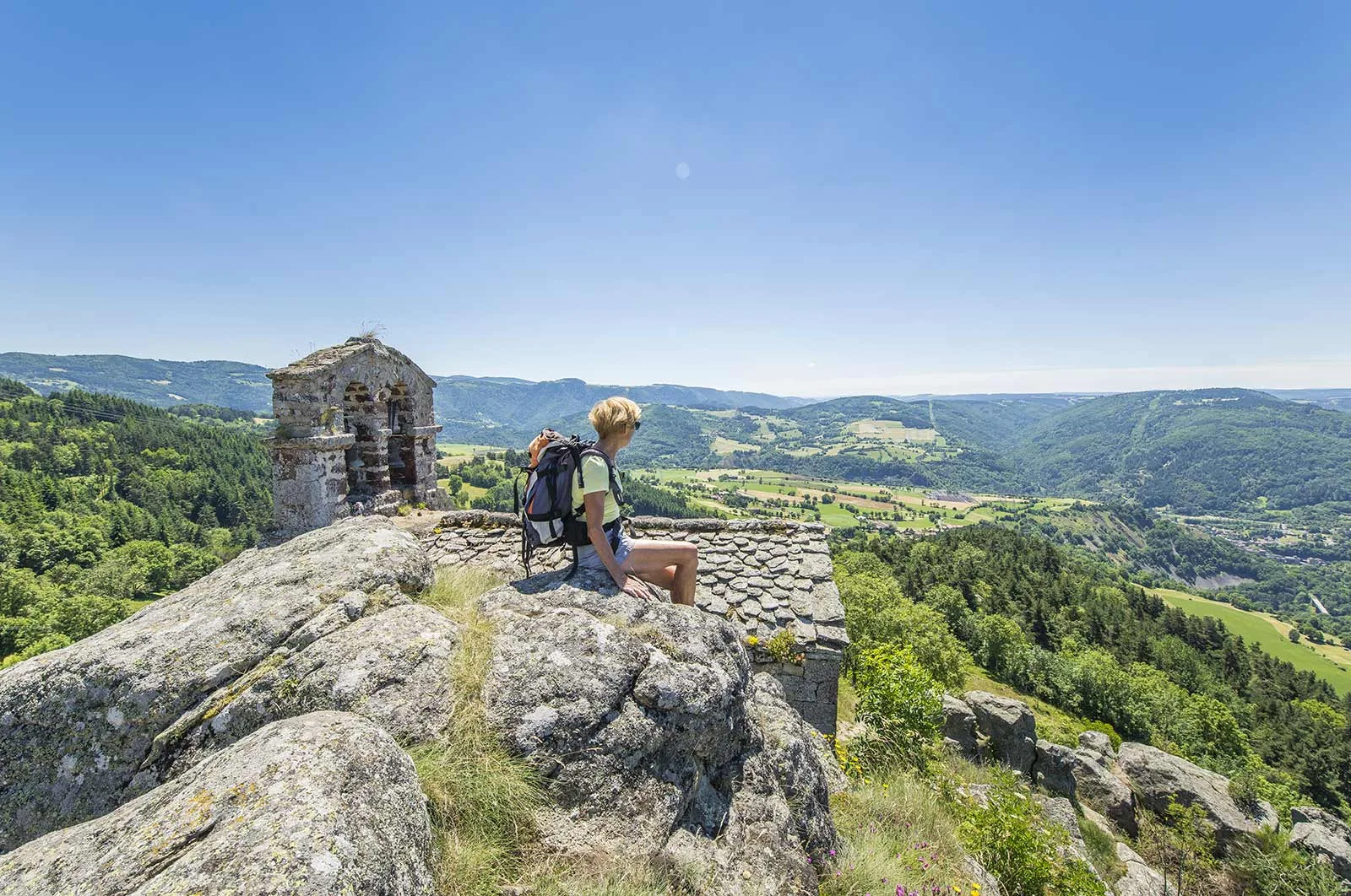 A woman sits on a rock and contemplates the sunny valley, way of Saint Jacques de Compostela (GR65) in Haute-Loire, Auvergne