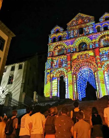 The crowd contemplates the Notre-Dame-du-Puy cathedral illuminated by the Puy en Lumières