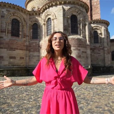 A young woman smiles with open arms in front of the Basilica of Saint-Julien de Brioude in Haute-Loire, Auvergne
