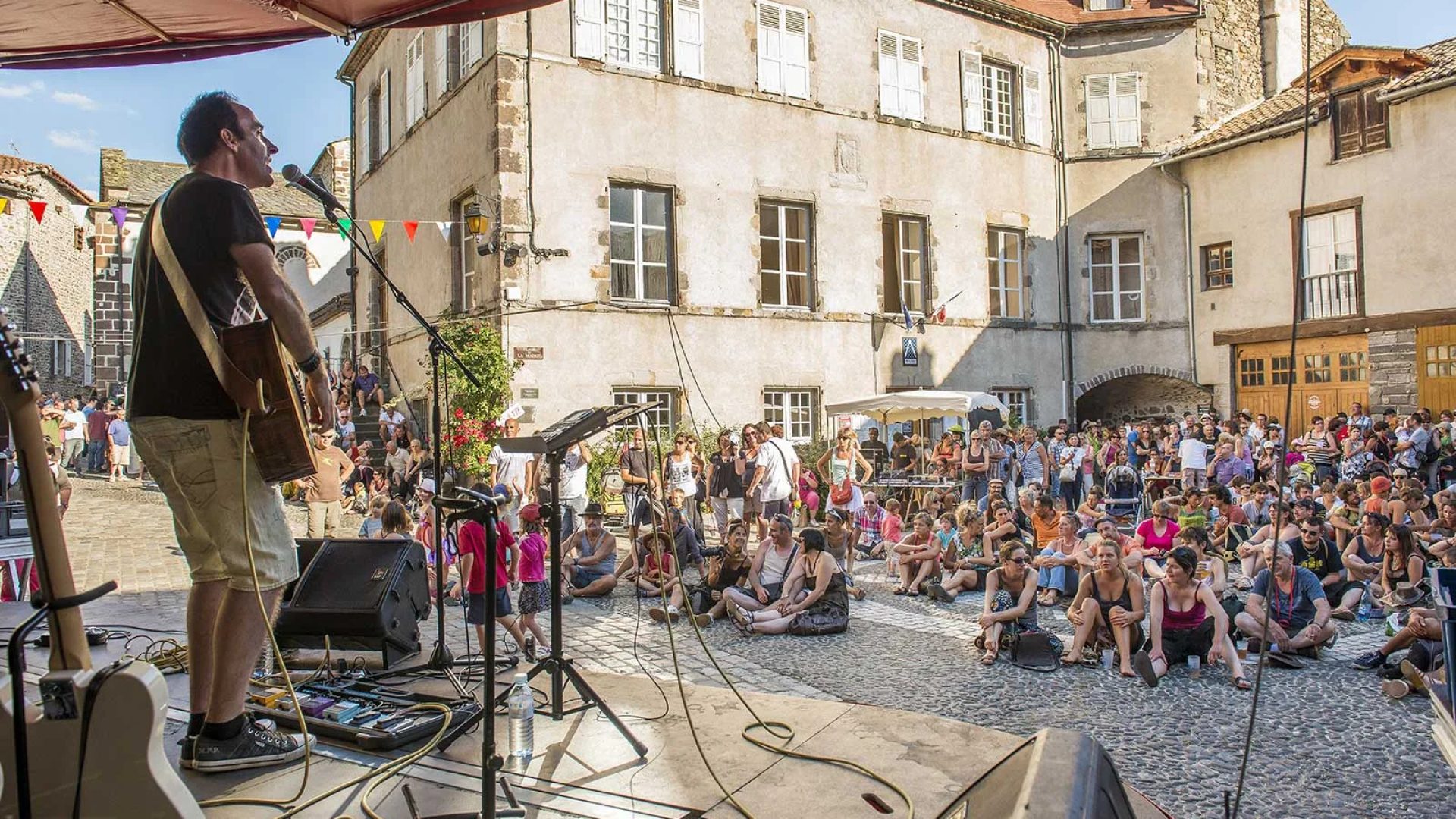Un cantante actúa en un escenario al aire libre frente a una multitud de personas sentadas en el Apéros musique de Blesle en Haute-Loire, Auvernia.