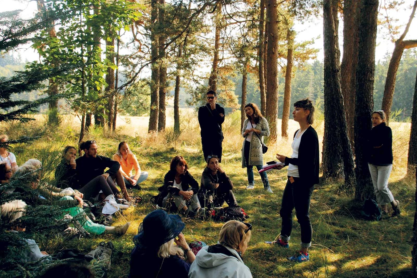 Un groupe de personnes est assis dans la forêt et écoute une femme compter une histoire pour les Lectures sous l'arbre en Haute-Loire, Auvergne