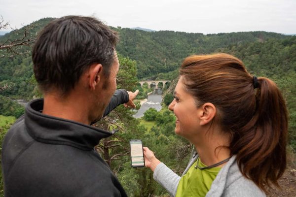 Un couple heureux regarde la vallée en direction d'un pont tout en suivant les indications de l'application Rando en Haute-Loire, Auvergne