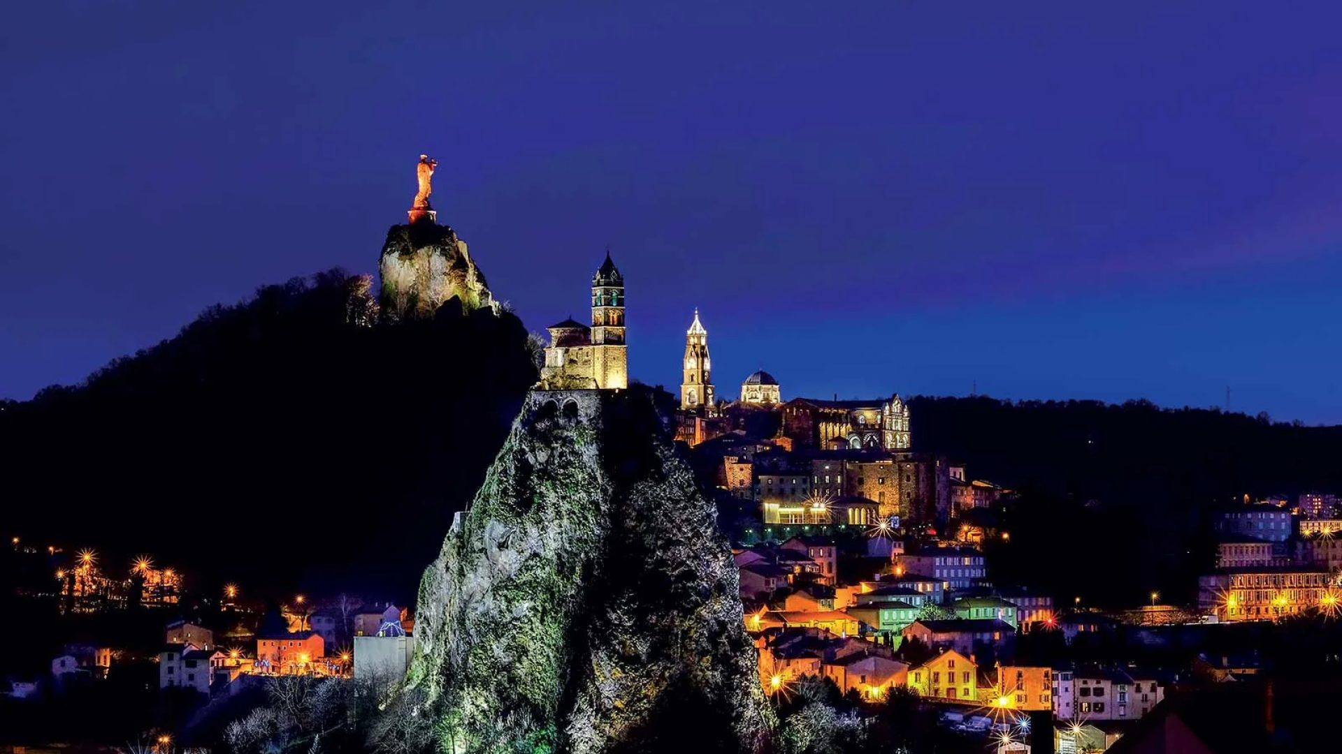 Nachtzicht op de Rocher Saint-Michel, het standbeeld van Notre-Dame de France en de stad Puy-en-Velay